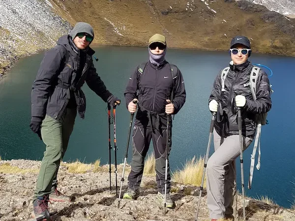 Three hikers with walking sticks and backpacks by a lagoon in the Cordillera Blanca near Huaraz, with snow-capped peaks in the background